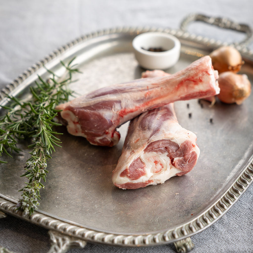 Raw Mount Bolton Springs Australian White lamb shank bones on a silver tray with herbs and a small bowl on a gray surface.