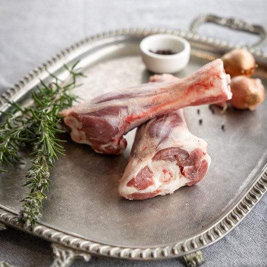 Raw Mount Bolton Springs Australian White lamb shank bones on a silver tray with herbs and a small bowl on a gray surface.